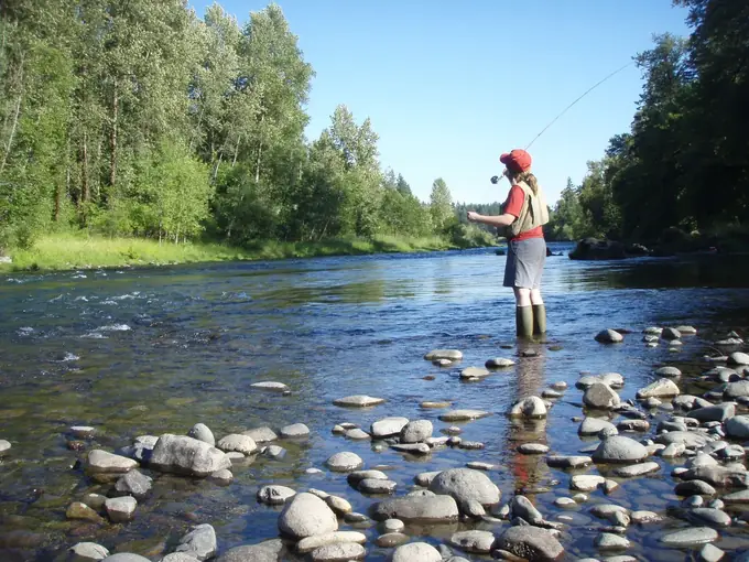 Pescadora com boné vermelho a lançar a linha num rio de pedras, entre árvores verdes